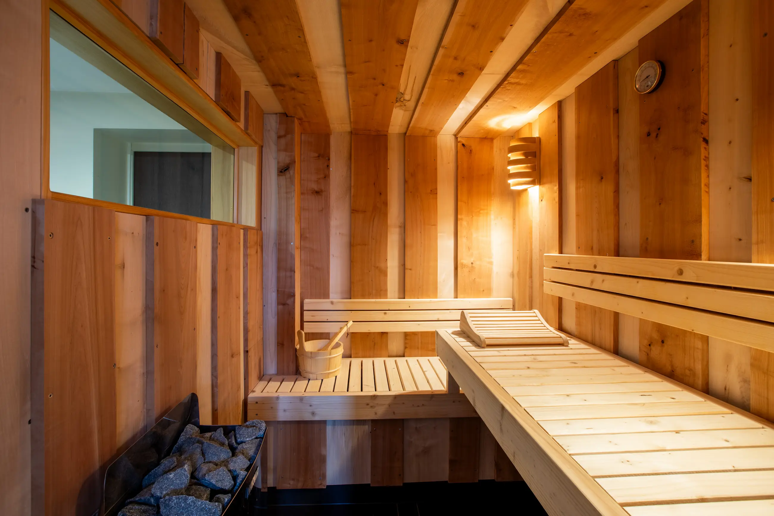 Sauna du gîte Porte D avec banquettes en bois, pierres volcaniques et lumière chaleureuse.